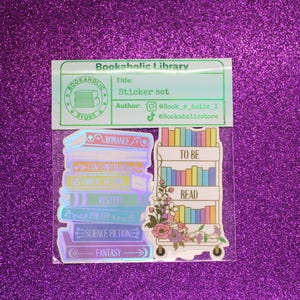 May include: A sticker set featuring book-themed designs. The set includes stickers of stacked books labeled with genres like "Romance" and "Fantasy," and a bookshelf on wheels with the text "To Be Read."