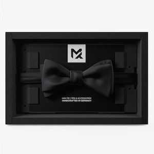 Może przedstawiać: Czarna mucha w czarnym pudełku. Pudełko ma zagłębienie, aby utrzymać muchę na miejscu. Na pudełku wydrukowano tekst "MAY-TIE | TIES & ACCESSORIES HANDCRAFTED IN GERMANY".