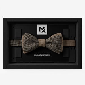 May include: A brown and black houndstooth bow tie and strap, displayed in a black presentation box. The bow tie has a classic pattern. The box includes the text "MAY-TIE | TIES & ACCESSORIES HANDCRAFTED IN GERMANY".