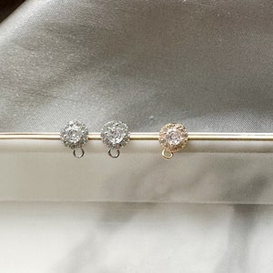 May include: Three stud earrings with a floral design, one gold-toned and two silver-toned, are displayed on a gold bar. A single gold earring is shown separately. The earrings have a central stone surrounded by smaller stones.