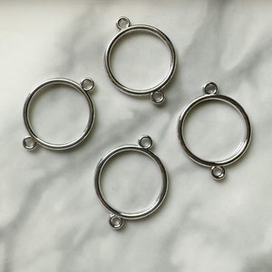 May include: Four silver metal rings with a circular shape and two small loops on each ring. The rings are arranged in a square shape on a white marble surface.
