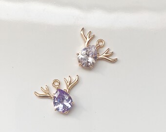 May include: Two gold-tone deer charms with crystal accents. The deer have antlers and a teardrop-shaped crystal in the center. One crystal is clear and the other is purple.