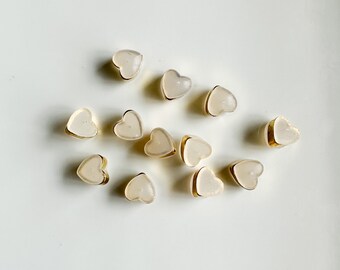 May include: A collection of small, translucent, heart-shaped charms with gold trim. The charms are scattered on a white surface.