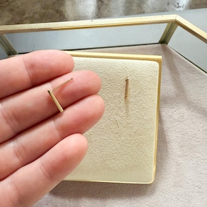 May include: A pair of gold-colored bar earrings. One earring is held in a hand, while the other is displayed on a cream-colored surface. The earrings have a minimalist design, with a straight bar and a post for piercing.