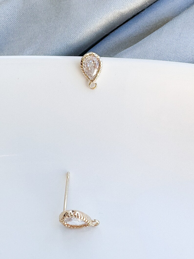 May include: A pair of gold teardrop shaped earrings with clear cubic zirconia stones. The earrings have a beaded edge and a small loop for hanging.