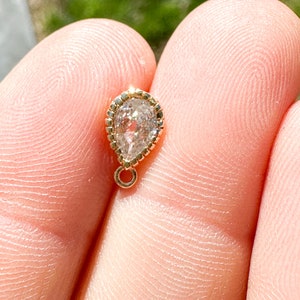May include: A small, teardrop-shaped pendant with a clear, faceted gemstone set in a gold bezel. The pendant has a delicate, gold chain attached to the top.
