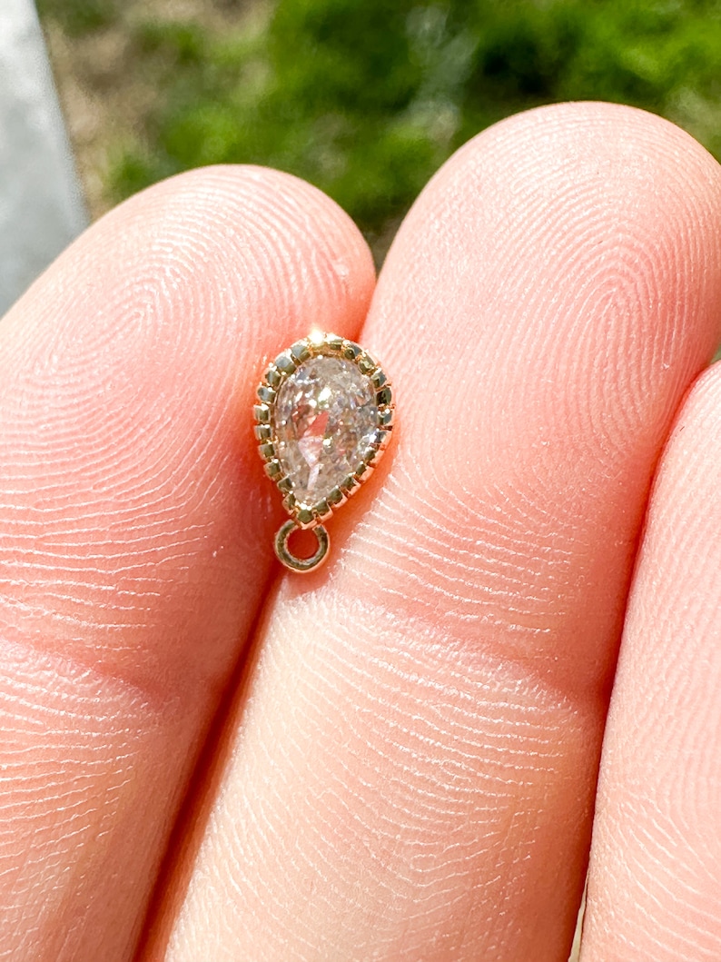 May include: A small, teardrop-shaped pendant with a clear, faceted gemstone set in a gold bezel. The pendant has a delicate, gold chain attached to the top.