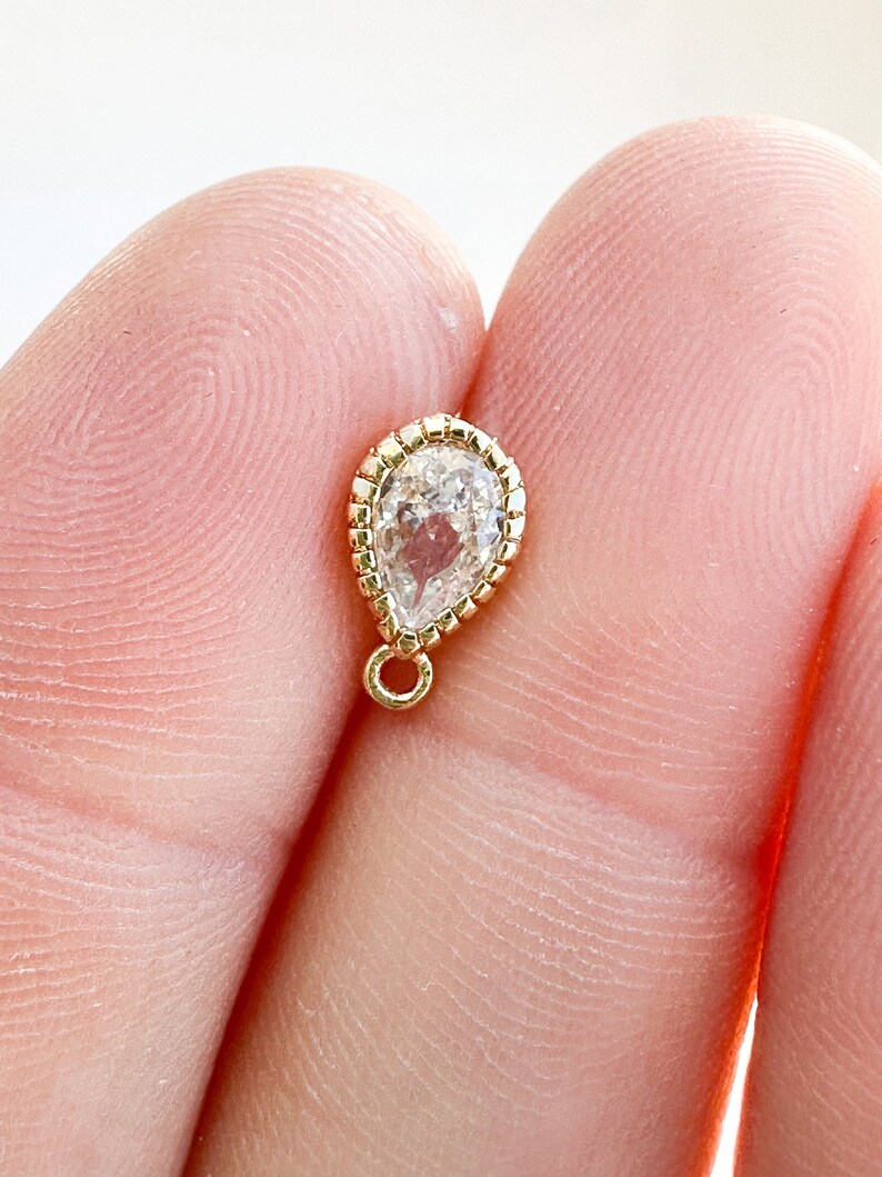 May include: A gold teardrop charm with a clear faceted gemstone and a small loop at the top.