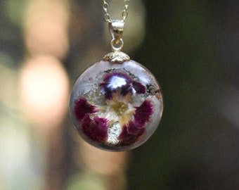May include: A gold chain necklace with a clear glass pendant containing a pressed purple and white flower.