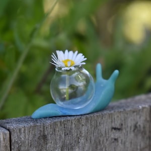 May include: A small blue snail figurine with a clear glass vase on its back, holding a single white daisy.