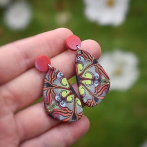 May include: A pair of teardrop-shaped earrings with a colorful, abstract design. The earrings are made of polymer clay and have red accents.