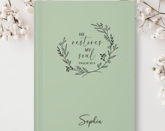May include: A sage green journal with a floral wreath design and the text "He restores my soul, Psalm 23:3". The name "Sophia" is written in cursive at the bottom.