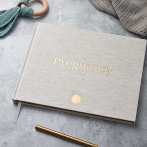 May include: A white linen-covered journal with gold lettering that reads "My Pregnancy Journal".