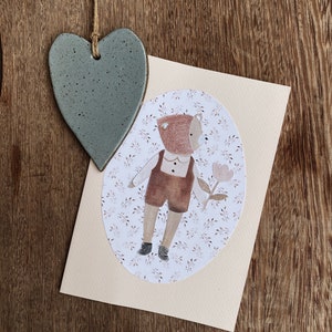 Puede incluir: Un adorno de coraz&oacute;n de cer&aacute;mica azul claro con una cuerda de yute marr&oacute;n colgando de &eacute;l. El adorno est&aacute; colgado sobre una tarjeta beige claro con una ilustraci&oacute;n de un oso marr&oacute;n sosteniendo una flor rosa. El oso lleva un sombrero marr&oacute;n y un mono marr&oacute;n.