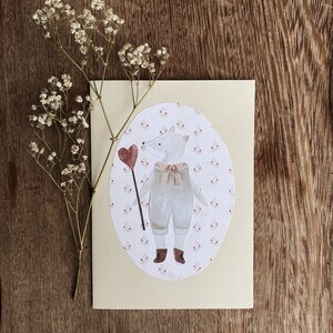 Puede incluir: Una tarjeta de color crema con un &oacute;valo blanco que presenta un zorro de dibujos animados que lleva un atuendo azul y sostiene un coraz&oacute;n rojo en un palo. El &oacute;valo est&aacute; rodeado por un peque&ntilde;o patr&oacute;n floral rosa.