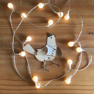 Puede incluir: Un p&aacute;jaro blanco con detalles marrones y un sombrero de copa negro est&aacute; rodeado de una cadena de luces blancas con bombillas transparentes. El p&aacute;jaro est&aacute; hecho de papel y tiene un aspecto artesanal y caprichoso.