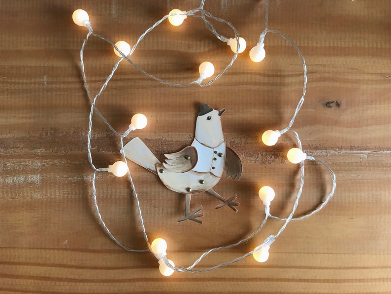 Puede incluir: Un p&aacute;jaro blanco con detalles marrones y un sombrero de copa negro est&aacute; rodeado de una cadena de luces blancas con bombillas transparentes. El p&aacute;jaro est&aacute; hecho de papel y tiene un aspecto artesanal y caprichoso.