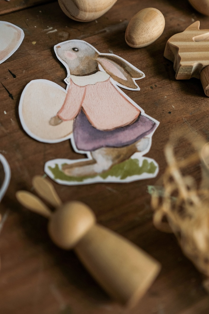 K&ouml;nnte beinhalten: Eine Aquarell-Illustration eines braunen Kaninchens, das ein wei&szlig;es Ei h&auml;lt. Das Kaninchen tr&auml;gt ein rosa Hemd und einen lila Rock. Die Illustration ist auf wei&szlig;em Hintergrund.