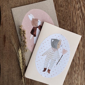 Puede incluir: Dos tarjetas de felicitaci&oacute;n con animales ilustrados. La primera tarjeta presenta un conejo marr&oacute;n que lleva un abrigo rojo y un sombrero rosa. La segunda tarjeta presenta un oso gris que lleva un mono a rayas y sostiene un globo rosa en forma de coraz&oacute;n.