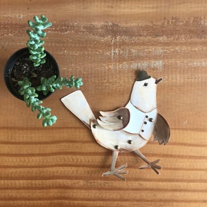 Puede incluir: Un p&aacute;jaro de papel blanco con detalles marrones y un sombrero de copa negro. El p&aacute;jaro lleva una camisa blanca con botones y est&aacute; de pie sobre una superficie de madera.