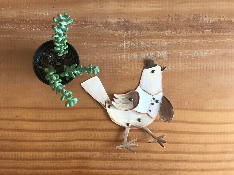 Puede incluir: Un p&aacute;jaro de papel blanco con detalles marrones y un sombrero de copa negro. El p&aacute;jaro lleva una camisa blanca con botones y est&aacute; de pie sobre una superficie de madera.
