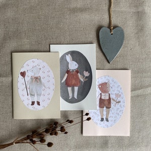 Puede incluir: Tres tarjetas de felicitaci&oacute;n ilustradas con un conejo, un zorro y un oso que llevan ropa y sostienen flores. Las tarjetas est&aacute;n sobre un fondo de lino beige con un adorno de coraz&oacute;n gris.