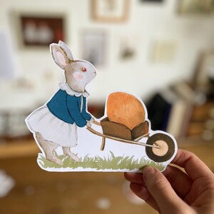 K&ouml;nnte beinhalten: Ein wei&szlig;es Kaninchen mit einer blauen Jacke und einem wei&szlig;en Rock schiebt eine Schubkarre mit einem gro&szlig;en orangen Gegenstand darin. Das Kaninchen steht auf gr&uuml;nem Gras.