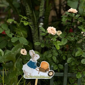 K&ouml;nnte beinhalten: Ein wei&szlig;es Kaninchen in einem blauen Kleid und einer wei&szlig;en Sch&uuml;rze schiebt eine Schubkarre mit einem gro&szlig;en orangefarbenen K&uuml;rbis darin. Das Kaninchen steht vor einem gr&uuml;nen Zaun und einem Busch mit rosa Bl&uuml;ten.