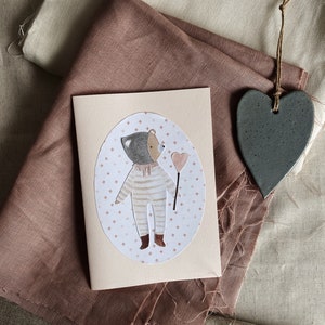 Puede incluir: Una tarjeta rosa con una ilustraci&oacute;n ovalada blanca de un oso con un mono de rayas y sosteniendo un coraz&oacute;n rosa. El oso est&aacute; de pie sobre un fondo de peque&ntilde;as estrellas blancas. La tarjeta est&aacute; sobre una tela marr&oacute;n.