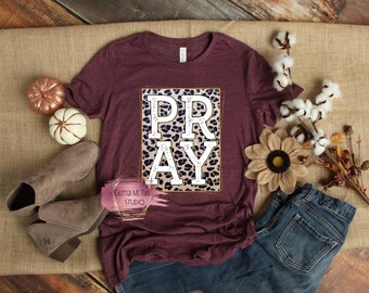 May include: A maroon t-shirt with a white "PRAY" graphic on a leopard print background. The graphic is surrounded by a gold outline.