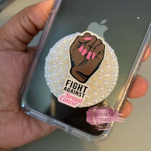 Puede incluir: Un soporte para teléfono blanco con un diseño de puño levantado y esmalte de uñas rosa. El texto "FIGHT AGAINST Breast Cancer" está en el soporte.