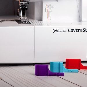 Peut inclure: Machine à coudre blanche Pacesetter Cover Stitch 2340CV avec trois guides de couture imprimés en 3D colorés. Les guides sont violets, bleus et rouges, avec le guide rouge étiqueté "1/2".
