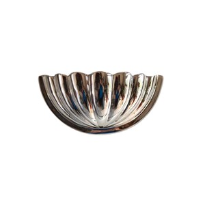 May include: A silver-colored, half-circle shaped decorative bowl with a fluted design. The bowl has a reflective, metallic finish and is set against a white background. The bowl's design features vertical, curved ridges that create a scalloped edge.