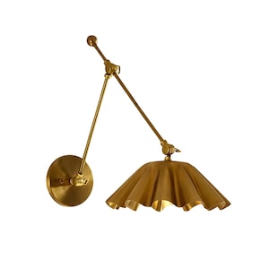 May include: A brass-toned wall sconce with an adjustable arm and a pleated lampshade. The round wall mount and the arm have a brushed finish. The lampshade has a ruffled, scalloped edge, and the light fixture is set against a white background.