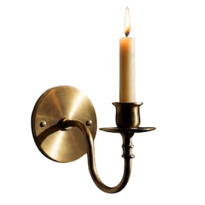 May include: A wall-mounted brass candle sconce with a circular backplate secured by two screws. The curved arm supports a candle cup and a drip tray. A single off-white taper candle is inserted into the cup, featuring a lit flame at the top.