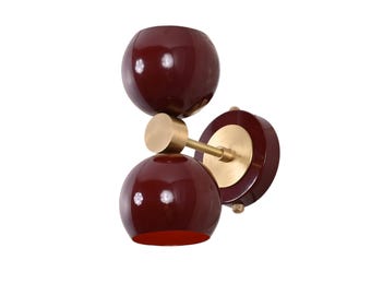 May include: A modern wall sconce featuring three burgundy-colored, spherical shades. The shades are connected by a brass-colored arm and mounted on a circular base. The sconce is designed for indoor lighting and has a contemporary aesthetic.
