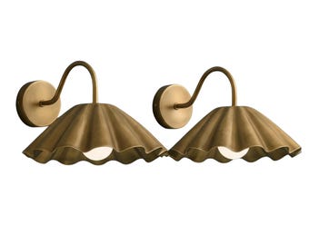 May include: A pair of wall-mounted light fixtures featuring a brushed brass finish. Each lamp has a circular wall plate, a curved arm, and a wide, scalloped, cone-shaped shade that directs light downward toward a single white bulb.