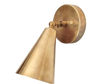 May include: A brass wall sconce with a conical shade and a circular backplate. The sconce is made of a shiny, golden-colored metal and is designed to be mounted on a wall. The shade is angled downward, and the fixture has a simple, modern design.
