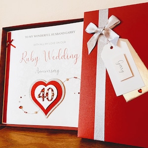May include: A red and white card with a heart-shaped design and the text "Ruby Wedding Anniversary" and "40". The card is in a red box with a silver ribbon and a white tag with the name "Garry" written on it.