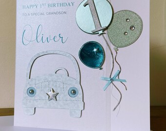 May include: A light blue card with the text "Happy 1st Birthday to a special grandson Oliver" and a silver number 1 balloon, two green balloons with silver glitter, a blue balloon with a silver star, and a white paper cut-out of a car.