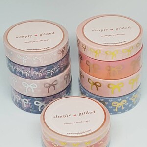 Può includere: Un set di rotoli di washi tape rosa e blu con fiocchi in lamina dorata. Il nastro è etichettato "simply gilded boutique washi tape" e include l'indirizzo del sito web "www.simplygilded.com".