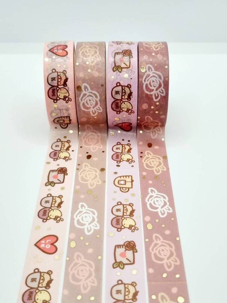 May include: Four rolls of washi tape with a pink and gold background. The tape features a repeating pattern of roses, gold glitter, and a couple of bears with the text "Be Mine".