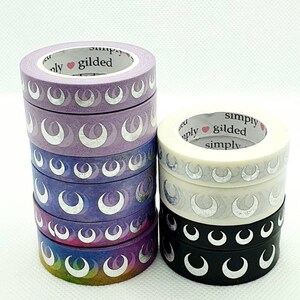 Può includere: Un rotolo di washi tape con un design a mezzaluna. I nastri sono in tonalità di viola, bianco e nero con accenti argentati. Il testo "simply gilded simply" è stampato sui nastri.