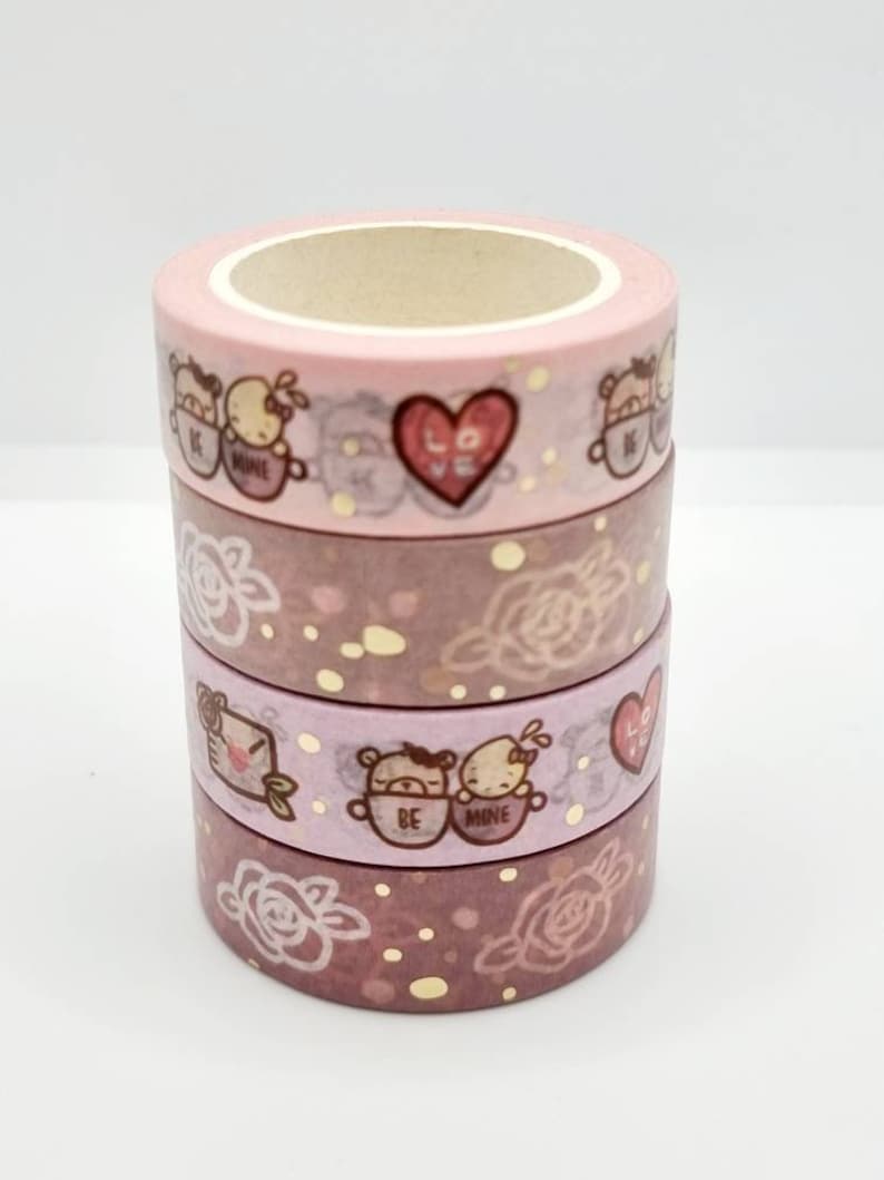 May include: A set of four pink and gold washi tapes with a romantic theme. The tapes feature illustrations of roses, hearts, and bears with the text "Be Mine" and "Love".