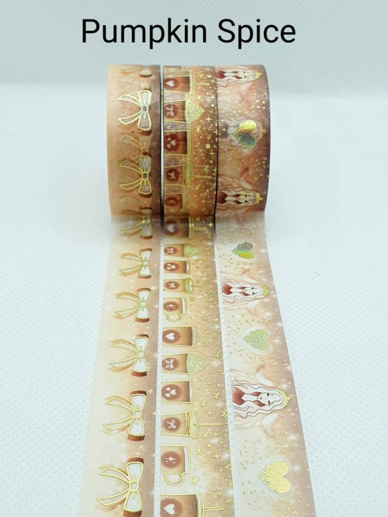 May include: Three rolls of washi tape with a fall theme. The tape features gold foil accents, illustrations of coffee cups, bows, and a woman with wings. The text "Pumpkin Spice" is at the top of the image.