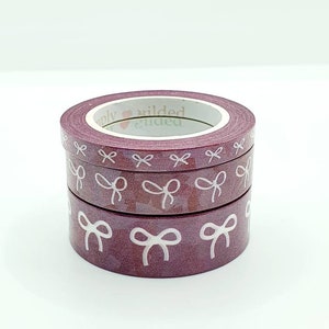 Pode incluir: Um rolo de fita washi com fundo bordô e desenhos de laços brancos. A fita é etiquetada como "Simply Wilded".