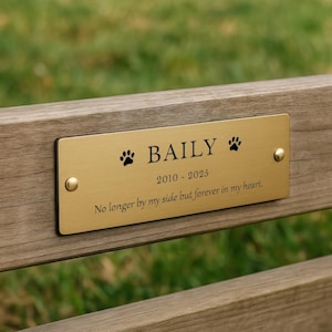May include: A gold-colored plaque with the name "BAILY" and paw prints is affixed to a wooden bench. The plaque includes the years 2010-2025 and the inscription "No longer by my side but forever in my heart."