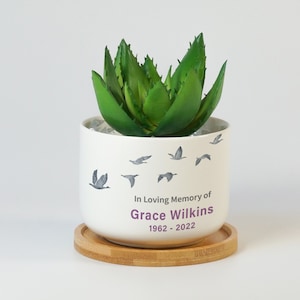 May include: White ceramic planter with a green succulent plant. The planter has a design of flying birds and the text "In Loving Memory of Grace Wilkins 1962 - 2022". The planter is sitting on a round wooden coaster.