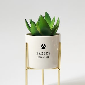 May include: White ceramic planter with a gold metal stand. The planter has a black paw print and the text "BAILEY 2010 - 2022".