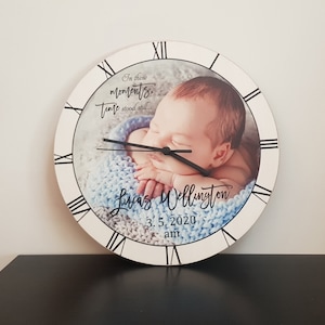 May include: A white wooden clock with a black face and Roman numerals. The clock has a photo of a baby sleeping on a blue blanket. The text "In these moments, time stood still..." is printed on the clock face. The baby's name, "Lucas Wellington", and the date and time of birth, "3.5.2020 am", are also printed on the clock face.